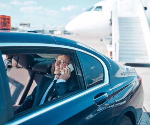Cheerful mature businessman is sitting in back of car and talking on cell phone while leaving airport after landing