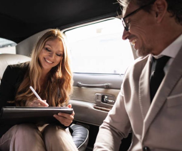 Businesswoman signing a contract on a digital tablet in the back of a limousine, with her business partner looking on